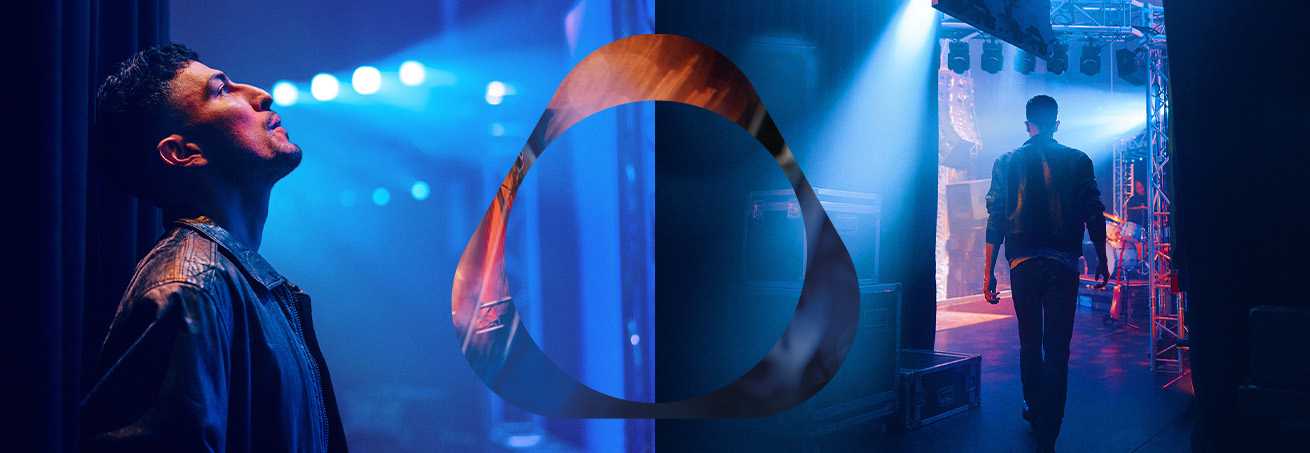 Cilvēks ādas jakā aizkulisēs zem zilām skatuves gaismām ar logo pārklājumu.