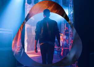 Cilvēks ādas jakā aizkulisēs zem zilām skatuves gaismām ar logo pārklājumu.