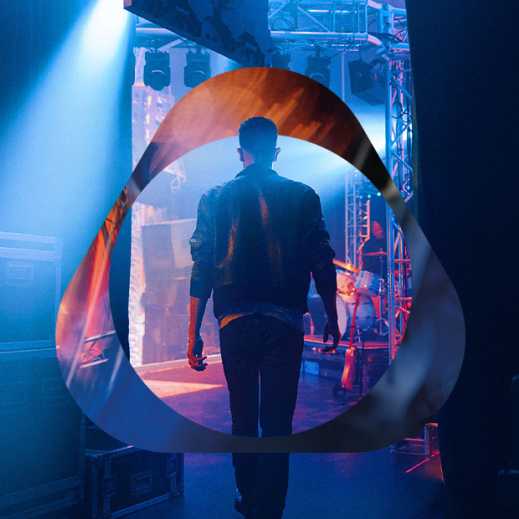 Cilvēks ādas jakā aizkulisēs zem zilām skatuves gaismām ar logo pārklājumu.