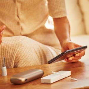 В руках телефон, на столе лежит IQOS ORIGINALS DUO.